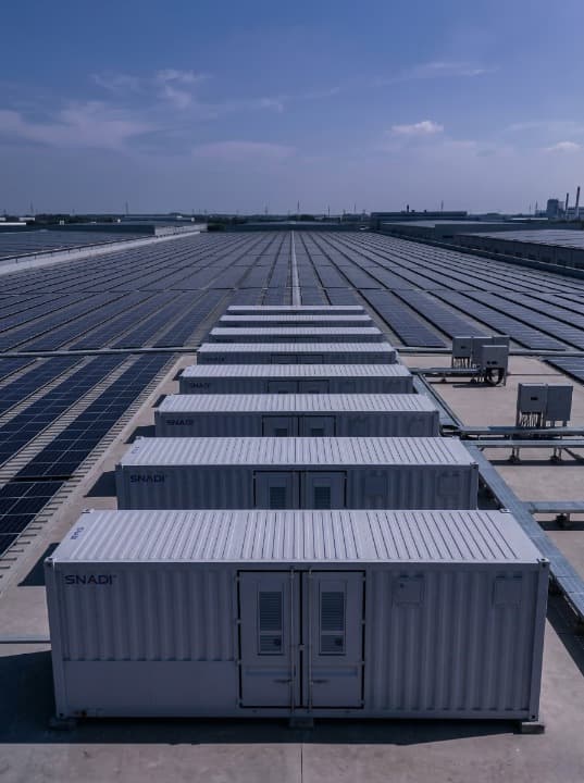 Una fila de gabinetes de almacenamiento de energía solar SNADI SNAT instalados frente a un enorme sistema solar fotovoltaico distribuido en la azotea en un complejo industrial de gran escala.